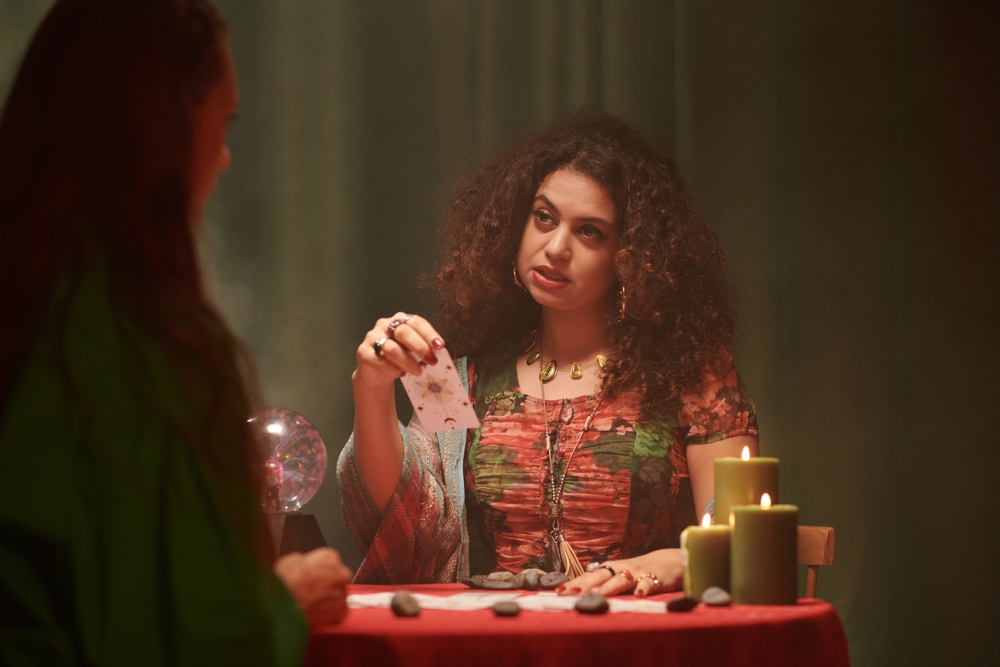 A tarot cards reader talking to a client. Both are sitting at a table with candles and a crystal ball.
